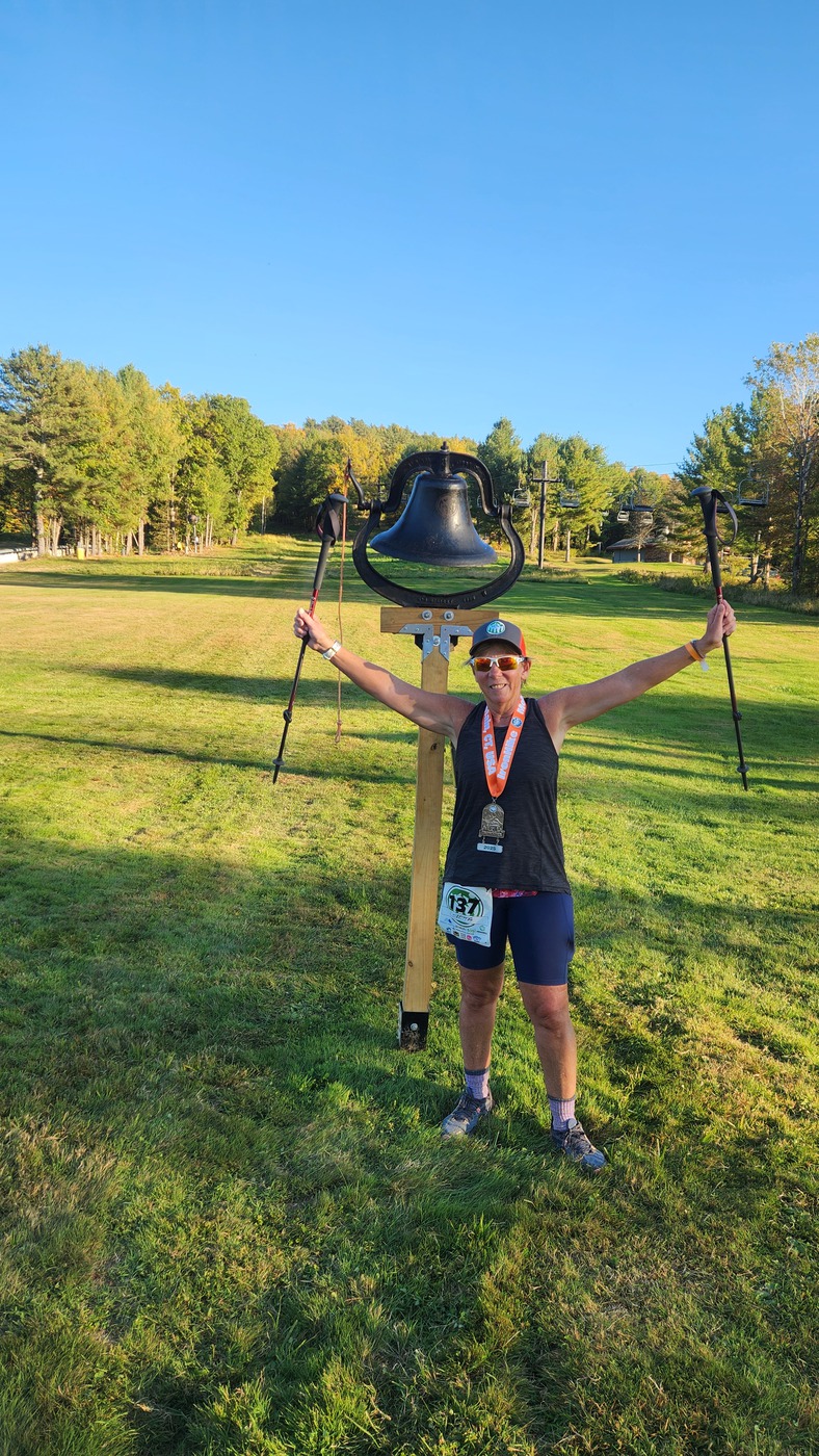 IronHike Finisher with arms and trekking poles raised and her medal at the bell, Mohawk Mountain